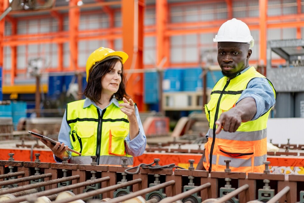 female supervisor advises technician foreman to inspect spare part equipment of production machine conveyor belt, team of diverse workers working together at industrial manufacturing factory
